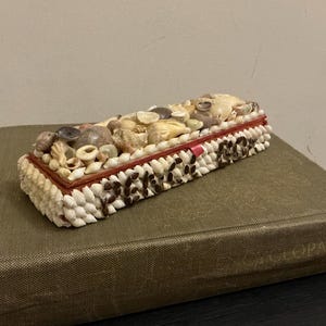 May include: A rectangular box covered in a mosaic of small seashells. The box is decorated with a red and white border and has a brown and white pattern on the bottom.