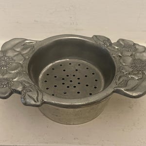 May include: A silver tea infuser with a floral design. The infuser has a strainer with small holes and two handles shaped like flowers.
