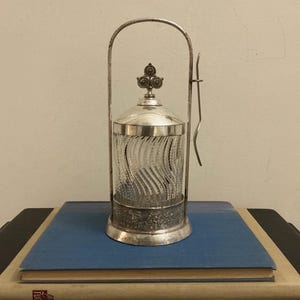 May include: A silver sugar bowl with a clear, textured glass container. The bowl has a decorative handle and a lid with a floral design. It sits on top of two books with blue and beige covers.