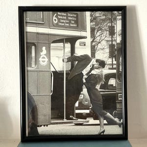 May include: Framed black and white photograph depicting a couple kissing near a London double-decker bus. The bus displays route information including 'West Kilburn' and 'Shoreditch'. A classic, romantic street scene.