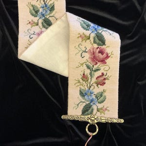 May include: An embroidered wall hanging with a floral pattern of pink roses and blue flowers. The beige fabric is detailed with green leaves and gold-toned metal bars at the top and bottom. A pink tassel hangs from the bottom.