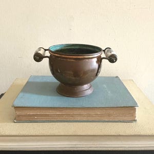 May include: A small, round copper bowl with a pedestal base and two side handles. The handles are made of white ceramic with black floral patterns. The interior of the bowl shows a green patina, while the exterior has a polished copper finish.