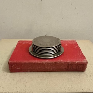 May include: A stack of six circular silver-toned metal coasters held within a matching metal stand. The top coaster features an intricate embossed floral pattern along its outer edge, while the remaining coasters have smooth, plain surfaces.