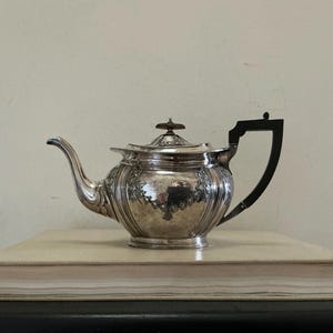 May include: A silver teapot with decorative floral patterns and a black handle. The teapot is placed on a beige book, with a light-colored wall behind it. The teapot features a curved spout and a small knob on the lid.