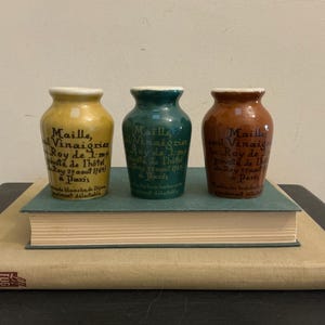 May include: Three small, antique ceramic jars in yellow, green, and brown, each with a white rim and French text. The jars are displayed on a stack of books with a light-coloured cover.