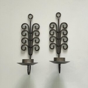 May include: A pair of dark grey metal wall sconces featuring a vertical stem with symmetrical, curling scrollwork branches and a circular ring at the top. Each sconce includes a flat, circular candle holder plate attached to the base.