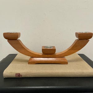 May include: A wooden candle holder with a curved design. The holder has three metal candle cups and is made of light brown wood. It sits on a beige book.