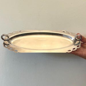May include: An oval silver-toned metal serving tray with a smooth, reflective central surface and a decorative scalloped edge. The tray features two small, circular ring-shaped handles attached to each end, providing a functional grip.