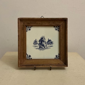 May include: A framed ceramic tile featuring a blue and white illustration of a painter at work, with a landscape scene. The tile is set in a square wooden frame with decorative corner details. The frame is supported by a small metal stand.