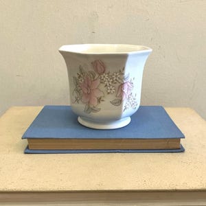 May include: A white ceramic planter with a hexagonal shape and a flared rim. The exterior features a delicate floral pattern in soft shades of pink, green, and grey, depicting clusters of flowers and leaves arranged around the centre of the vessel.