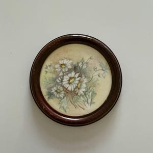 May include: A small, circular painting in a dark brown wooden frame. The artwork depicts a cluster of white daisies with yellow centres and green foliage, set against a pale yellow backdrop.