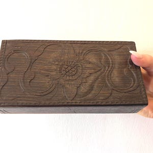 May include: A rectangular, dark brown wooden box with a carved floral design on the lid. The box is held by a hand. The box is likely used for storing small items.