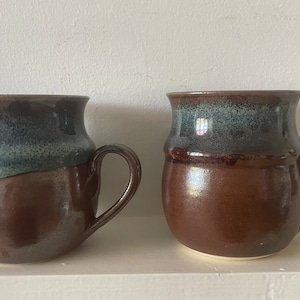 May include: Two brown ceramic mugs with a blue and green glaze. The mugs have a rounded body and a handle.