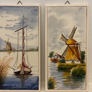 May include: Two vintage Dutch tile paintings. The first tile depicts a sailboat with a red sail on a canal with a windmill in the background. The second tile depicts a yellow windmill with a white house on a canal with green reeds.