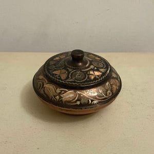May include: A round metal lidded container featuring a dark brown finish with intricate copper and silver-toned floral engravings. The lid has a central knob and a scalloped border, while the body displays detailed tulip and botanical patterns.