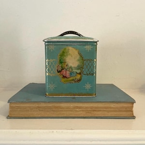 May include: A vintage teal metal tin with a handle, featuring a pastoral scene on the front. The tin sits on a light blue book. The tin has gold accents and a decorative design.