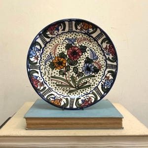 May include: A decorative ceramic plate with a hand-painted floral design. The plate has a dark blue rim and a white background with colorful flowers in red, orange, and blue. The plate is approximately 25 cm in diameter.