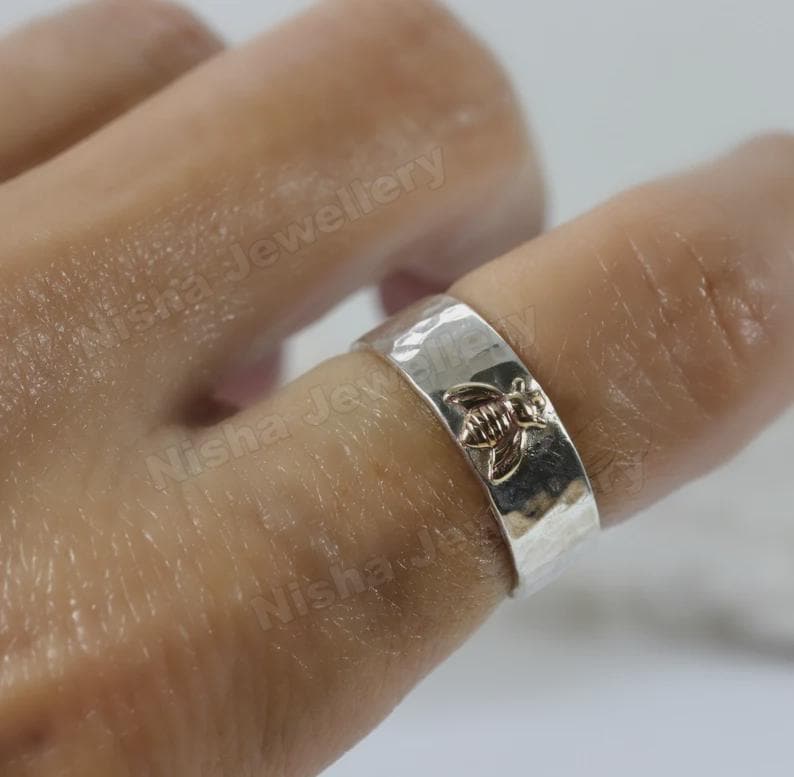 May include: A silver ring with a hammered texture and a raised bee design. The ring is worn on a finger, showcasing the detailed craftsmanship of the jewelry. The ring is a wide band style.