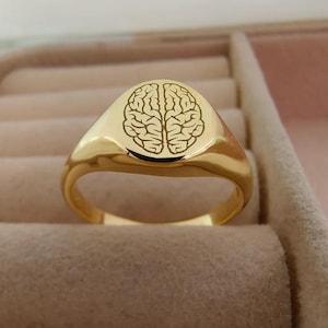 May include: A gold-colored signet ring with a detailed brain design engraved on the oval face. The ring has a polished, reflective surface and a classic, rounded band. The ring is displayed in a jewelry box.