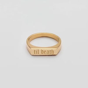 以下が含まれることがあります： ゴシック体で「til death」と刻まれたゴールドカラーのリング。 リングは長方形の表面と滑らかで磨かれたバンドを備えています。 リングは無地の白い背景に配置されています。