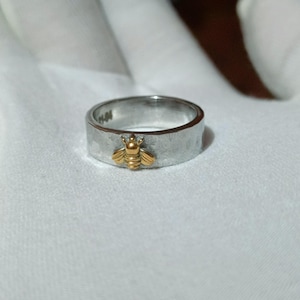 May include: A silver ring with a textured band and a gold-colored bee charm. The bee is centered on the ring and has detailed wings and body. The ring is shown on a white surface.
