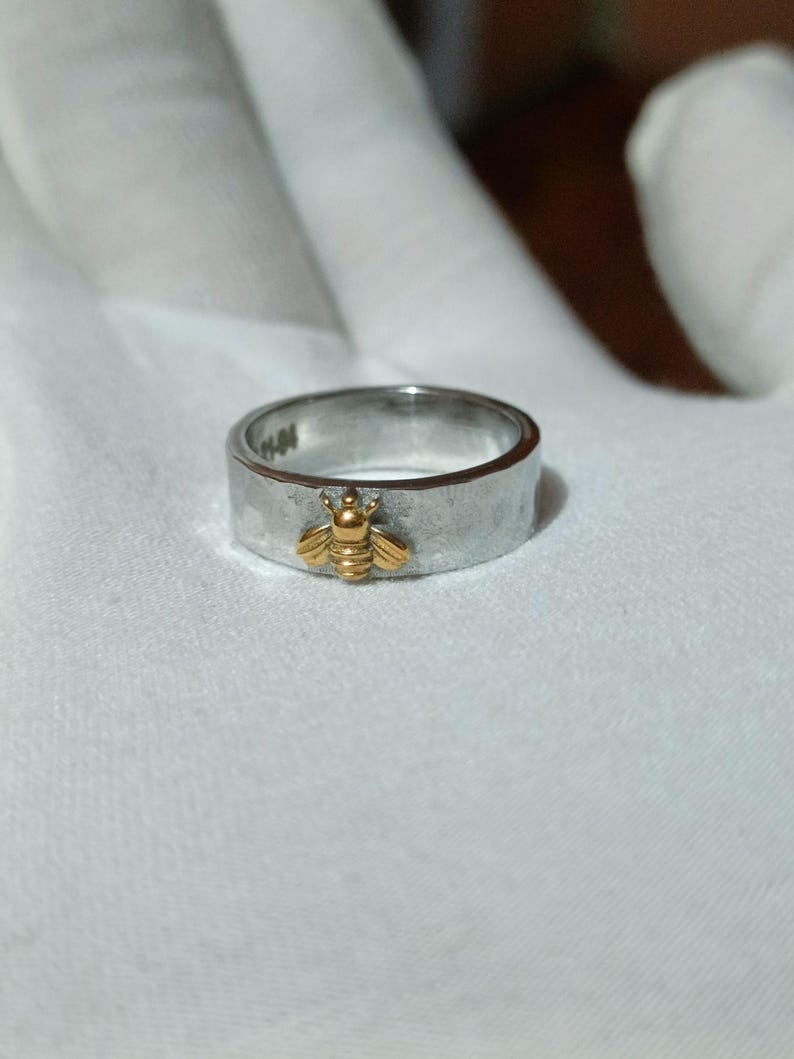 May include: A silver ring with a textured band and a gold-colored bee charm. The bee is centered on the ring and has detailed wings and body. The ring is shown on a white surface.