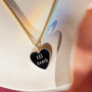 Puede incluir: Un collar de cadena dorada con un colgante de corazón negro. El colgante de corazón tiene un borde plateado y las palabras "til death" en letras góticas blancas. El collar se muestra sobre una superficie blanca.