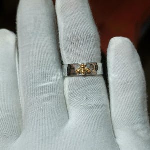 May include: A silver ring with a hammered texture and a gold-colored bee design. The bee is centered on the ring, which is shown on a finger.