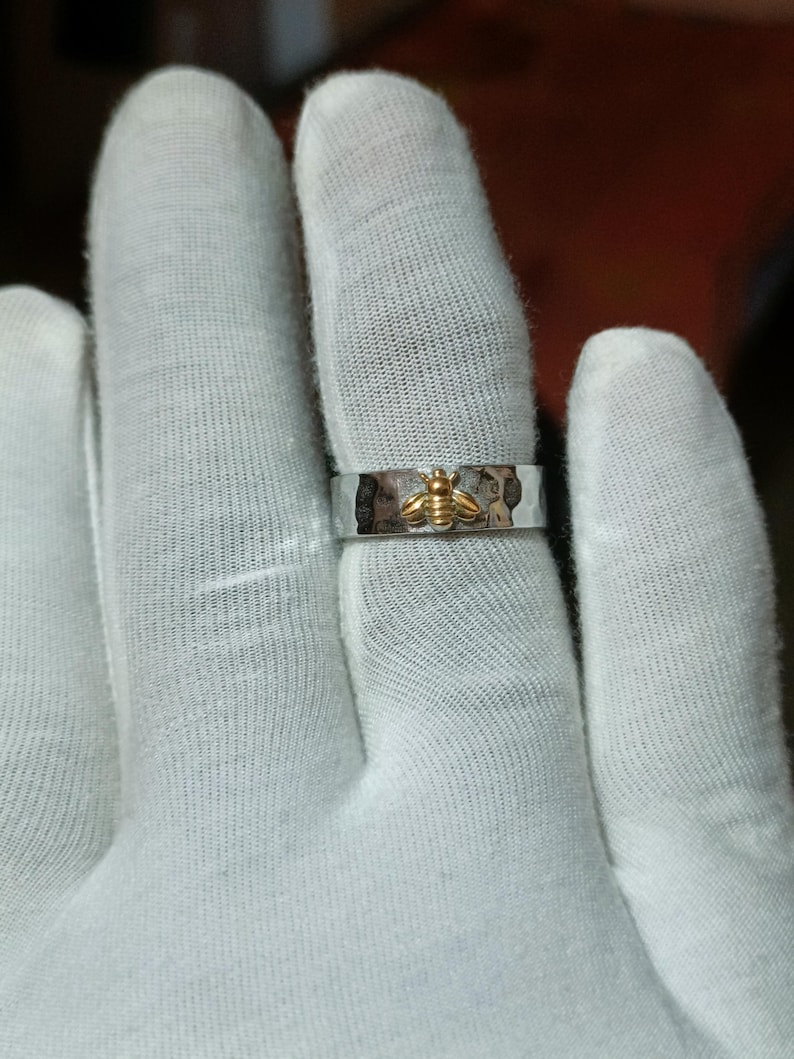 May include: A silver ring with a hammered texture and a gold-colored bee design. The bee is centered on the ring, which is shown on a finger.
