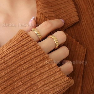 Può includere: Due anelli dorati con un design intrecciato strutturato. Gli anelli sono indossati sulle dita, su uno sfondo marrone caldo. Gli anelli sono il punto focale, mostrando uno stile di gioielleria classico.