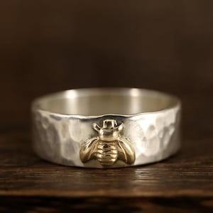May include: A silver ring with a hammered texture and a gold-colored bee emblem. The ring is wide and sits on a wooden surface. The bee is detailed with wings and body segments.