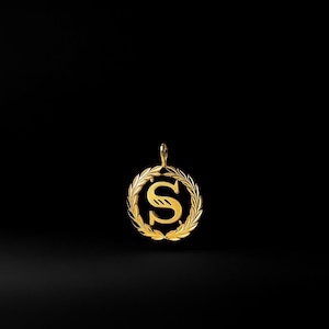 Könnte beinhalten: Goldfarbener Anhänger mit dem Buchstaben „S“ in dekorativer Schrift, umgeben von einem Lorbeerkranz. Der Anhänger hat eine Schlaufe für eine Kette und ist vor einem schwarzen Hintergrund platziert.