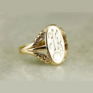 Op de afbeelding: Een goudkleurige zegelring met een ovale voorkant. De voorkant is gegraveerd met de letter "R" in een decoratief lettertype. De ring heeft sierlijke details op de schouders en een gepolijste afwerking.