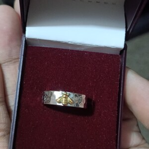 May include: A silver ring with a hammered texture and a gold-colored bee design. The ring is displayed in a burgundy jewelry box with a white interior.