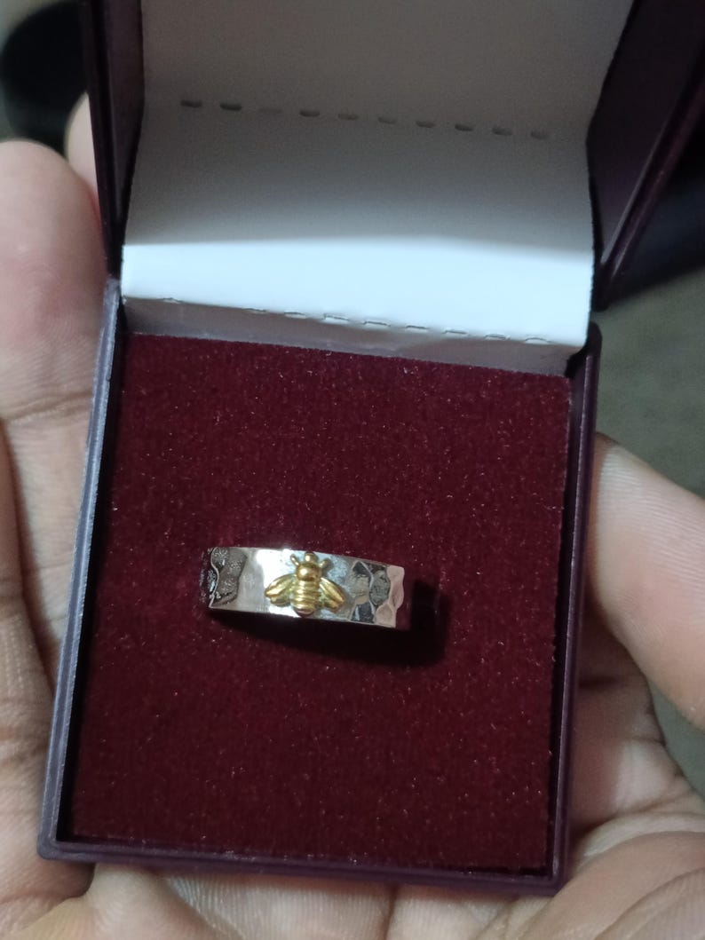 May include: A silver ring with a hammered texture and a gold-colored bee design. The ring is displayed in a burgundy jewelry box with a white interior.