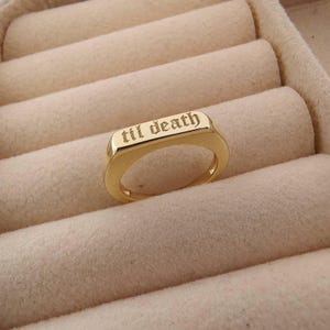 May include: A gold-colored ring with the inscription "til death" in a gothic font. The ring has a rectangular face and a smooth, polished finish. The ring is displayed on a beige, textured surface.
