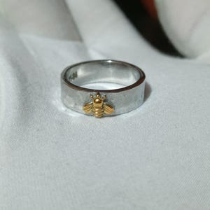 May include: A silver ring with a gold-colored bee charm. The ring has a brushed metal finish and a flat band. The bee is centered on the ring and has detailed wings and body. The ring is a simple, elegant piece of jewelry.