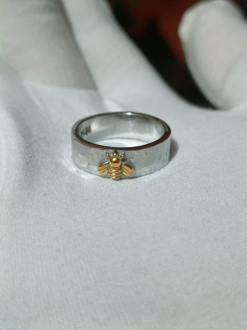 May include: A silver ring with a gold-colored bee charm. The ring has a brushed metal finish and a flat band. The bee is centered on the ring and has detailed wings and body. The ring is a simple, elegant piece of jewelry.