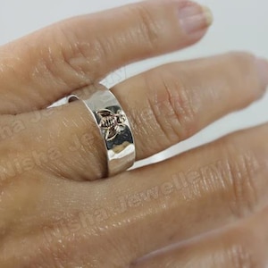 May include: A silver ring with a floral design. The ring is wide and has a hammered texture. The floral design is centered on the ring and is slightly raised. The ring is worn on a finger.