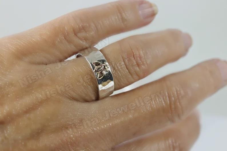 May include: A silver ring with a floral design. The ring is wide and has a hammered texture. The floral design is centered on the ring and is slightly raised. The ring is worn on a finger.