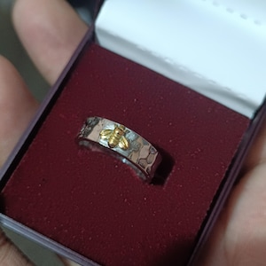 May include: A silver ring with a hammered texture and a gold bee design. The ring is presented in a burgundy jewelry box with a white interior. The bee is centered on the ring.