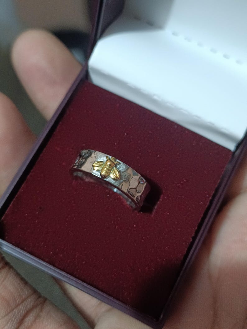 May include: A silver ring with a hammered texture and a gold bee design. The ring is presented in a burgundy jewelry box with a white interior. The bee is centered on the ring.