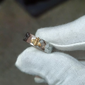 May include: A silver ring with a hammered texture and a gold-colored bee design. The bee is centered on the ring, which is held by a gloved hand. The ring is a simple band style.