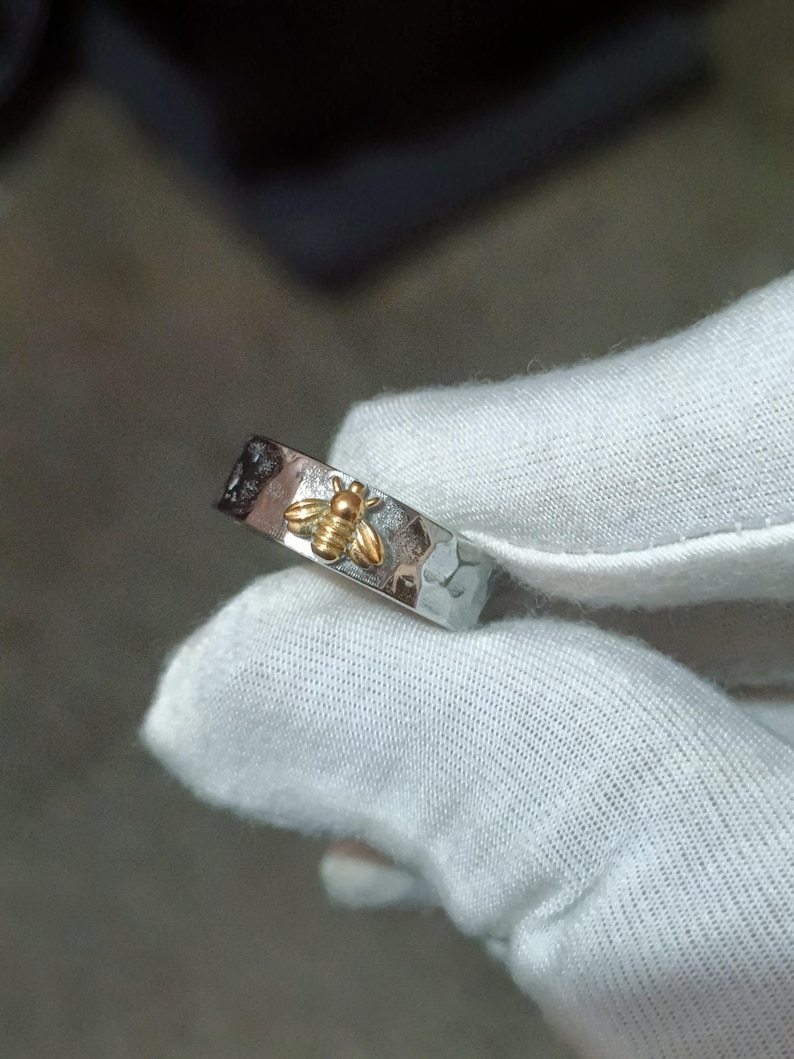 May include: A silver ring with a hammered texture and a gold-colored bee design. The bee is centered on the ring, which is held by a gloved hand. The ring is a simple band style.