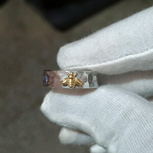 May include: A silver ring with a hammered texture and a gold bee design. The ring has a small blue gemstone and is held by a gloved hand. The bee is centered on the ring.