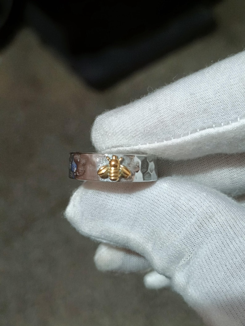 May include: A silver ring with a hammered texture and a gold bee design. The ring has a small blue gemstone and is held by a gloved hand. The bee is centered on the ring.