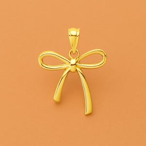 Könnte beinhalten: Ein goldfarbener Anhänger in Form einer Schleife. Die Schleife hat oben eine Schlaufe für eine Kette und zwei gebogene Enden. Der Anhänger ist vor einem orangefarbenen Hintergrund platziert.