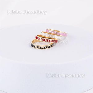 Puede incluir: Cuatro anillos de esmalte personalizados en oro. Cada anillo presenta un nombre diferente: Jennifer, Patricia, Linda y Mary. Los anillos tienen esmalte negro, rojo, blanco y rosa con texto a juego. Los anillos están apilados sobre una superficie blanca.
