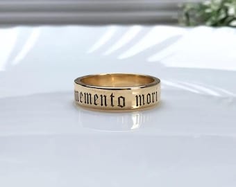 May include: A gold-colored ring with the words "memento mori" engraved in a gothic font. The ring is set against a white background, highlighting its polished surface and the dark lettering.