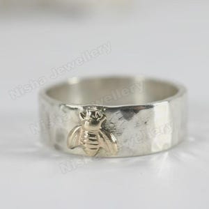 May include: A silver ring with a hammered texture and a gold-colored bee charm. The bee is centered on the ring and has detailed wings and body. The ring is a wide band style.
